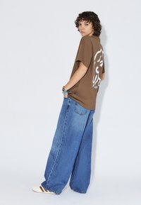 T-shirt marron oversize avec un imprimé graphique au dos, associé à un jean bleu à jambes larges. Détails comprenant une coupe décontractée et des manches retroussées.