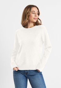Witte gebreide trui met geribbelde halslijn en boorden. Zachte textuur. Gecombineerd met blauwe denim jeans, wat de casual pasvorm benadrukt.