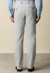 Lichtgrijze broek van een gestructureerde stof, met twee achterzakken met knopen, recht gesneden pijpen, en gedragen met een paar bruine schoenen.