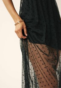 Schwarzes, durchsichtiges Kleid mit Polka-Dots, geschichteter Stoff und einem seitlichen Schlitz, der einen einfarbigen Ton darunter zeigt. Goldene Kettenarmband am Handgelenk.