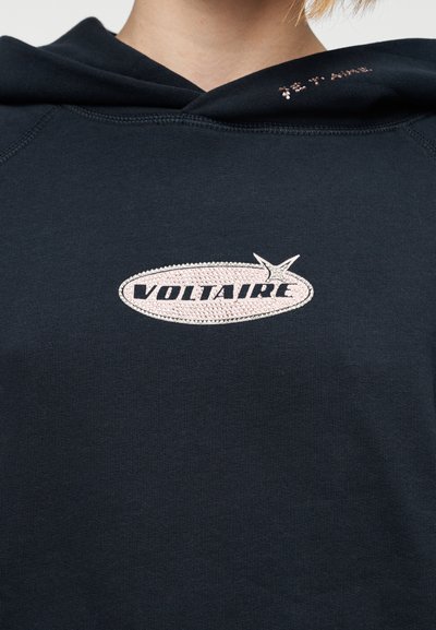 Φούτερ με κουκούλα σε ναυτικό μπλε χρώμα με οβάλ εφαρμογή που φέρει τη λέξη «VOLTAIRE» μαύρα, περιτριγυρισμένη από υφασμένο σχέδιο. Λεπτομέρειες διακριτικής έμφασης στην κουκούλα.