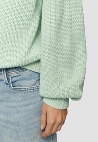 Maglione lavorato a maglia verde chiaro con pattern strutturato, maniche larghe e polsini a coste, indossato sopra jeans di denim blu chiaro.