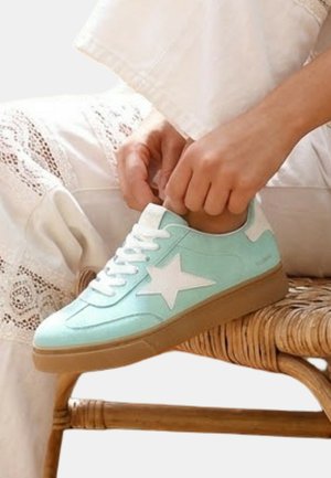 Personne portant un pantalon en dentelle blanche en train de nouer les lacets d'une basket bleu clair avec une étoile blanche, assise sur une chaise tressée.