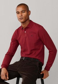 Polo à manches longues bordeaux avec col, arborant un petit logo sombre sur la poitrine. Associé à un pantalon en velours côtelé foncé et une ceinture noire.