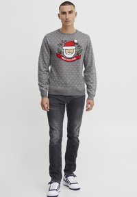 Pull gris avec un motif de Santa, le texte "HO HO HO" et des accents de houx verts. Présente de petits motifs blancs et des poignets côtelés au niveau du col.
