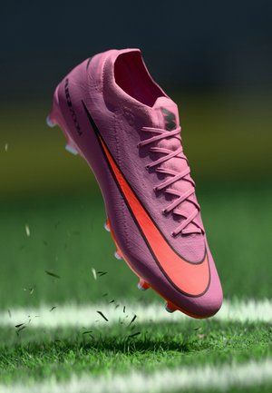 Roze en oranje voetbalschoen zwevend boven gras waarbij de noppen kleine stukjes gras op een gemarkeerd veld opstoten.