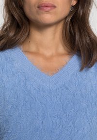 Ljusblå, fluffig stickad tröja med V-ringning och strukturerat kabelmönster. Mjuk vaddering med synliga fluffiga fibrer runt halsringningen och kroppen.