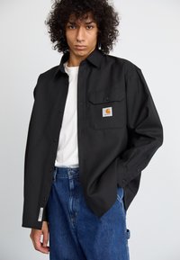 Fekete gombos inget visel, mellzsebes és Carhartt logóval, fehér póló felett, magas derekú kék farmernadrággal párosítva.