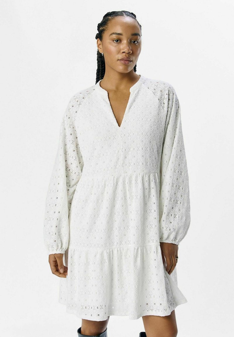 Robe blanche mi-longue avec de longues manches bouffantes. Le tissu en œillets présente des motifs en losange et un décolleté en V. Ourlet évasé.