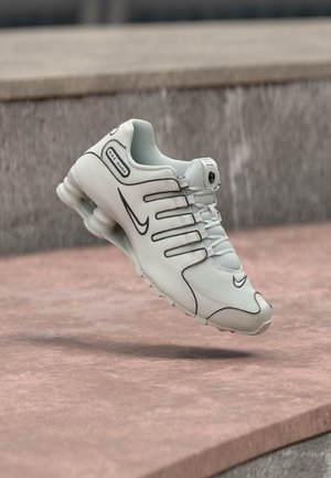 Biela teniska Nike Shox s čiernym logom a odpruženou podrážkou, umiestnená vo vzduchu nad ružovým betónovým povrchom.