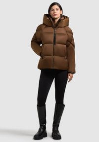 Giacca imbottita marrone con colletto alto e cappuccio, dotata di zip frontale. Il modello indossa leggings neri e stivali neri alti fino al ginocchio.