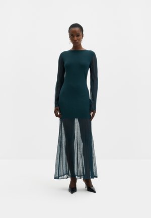 Femme portant une robe ajustée bleu sarcelle foncé avec des manches longues transparentes et une jupe longue transparente, associée à des talons pointus noirs.