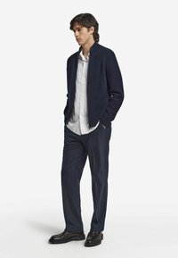 Veste bomber en maille côtelée marine, chemise blanche boutonnée, pantalon bleu marine et chaussures en cuir noir. Le modèle se tient les mains dans les poches.