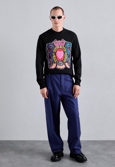 Svart stickad tröja med en färgglad grafisk design med motiv och ett hjärta. Kombinerad med lösa marinblå byxor och svarta skor.