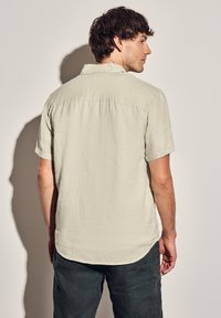 Camisa de manga corta verde clara hecha de tela texturizada, con dobladillo recto y un pequeño yugo trasero, mostrada desde la parte de atrás.