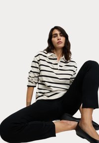 Camisa polo de manga larga a rayas blancas y negras, de corte relajado, combinada con pantalones negros y zapatos negros sin cordones, llevada por una persona sentada.