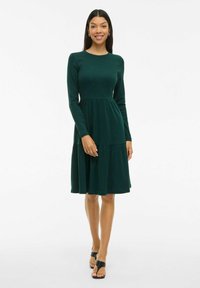 Robes longue à manches longues vert foncé avec un corsage ajusté et une jupe à volants. Fabriquée à partir d'un tissu doux, elle possède un col rond et un design simple.