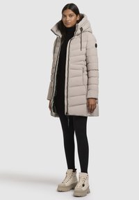 Manteau matelassé en beige clair, avec un col haut, un design matelassé horizontal et des poches zippées. Associé à des leggings noirs et des bottes épaisses beiges.