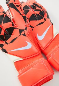 Nike rukavice za golmana u vibrantnoj narančastoj boji s geometrijskim crnim uzorkom. Imaju teksturirani grip, sigurnu narukvicu i bijele akcentne detalje.