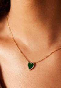 Gouden kettinghalsketting met een hartvormige groene steenpendant, omringd door kleine heldere kristallen, rustend op een gladde huid.