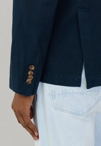 Manche d'un blazer bleu foncé avec quatre boutons marron sur une poche de jean bleu clair, main détendue le long du corps.