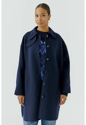 Vrouw draagt een marineblauwe jas met knopen over een donkerblauwe gebreide trui met patroon en lichtblauwe jeans, staand tegen een effen lichte achtergrond.