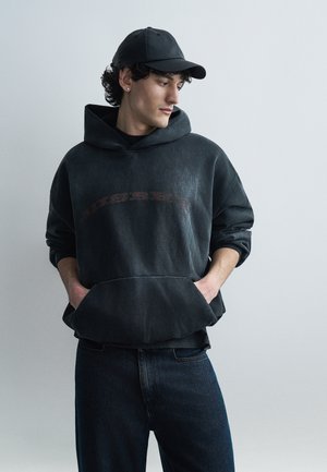 Mladenič z kodrastimi lasmi nosi črno kapo, izpran črn pulover s kapuco z rokami v žepih in temne kavbojke ter gleda na desno ob enostavni ozadju.