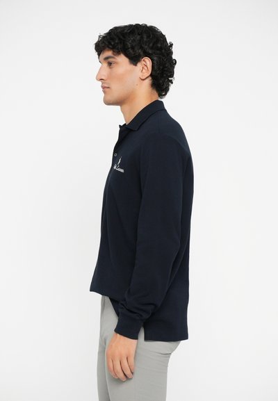 Camisa polo de manga larga azul marino con cuello y logo bordado, hecha de una tela suave. Modelo vistiendo pantalones gris claro.