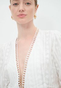 Weißes besticktes Kleid mit tiefem V-Ausschnitt, strukturiertem Blumenmuster und kontrastierendem, gewelltem Saum am Ausschnitt. Goldene geometrische Ohrringe.