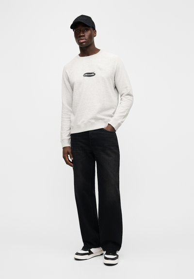 Light gray sweatshirt with a black logo, paired with loose black jeans and black-and-white sneakers. Cap with the brand name in white.