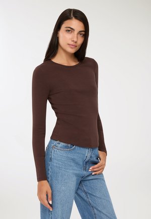 Camicia marrone a maniche lunghe realizzata in tessuto morbido, con scollatura rotonda e taglio aderente, abbinata a jeans a vita alta blu chiaro.