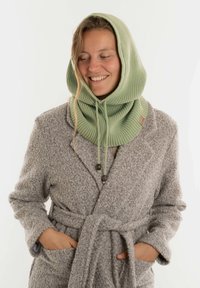 Groene gebreide capuchonsjaal met een geribbeld patroon en koordjes. G gedragen over een grijze gewatteerde rok met een gestrikte taille.