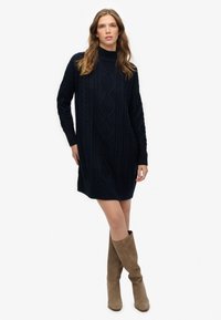 Vestido de punto azul marino con cuello alto, que presenta un patrón de trenzas texturizado, mangas largas y un ajuste relajado, combinado con botas hasta la rodilla color topo.