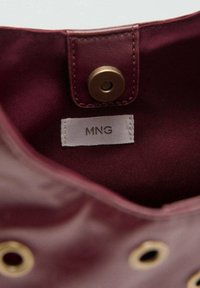 Viininpunainen tekstiilisaapas, jossa on kangasvuori valkoisella "MNG"-etiketillä ja kultaisen sävyinen metallinen nepparikiinnitys.