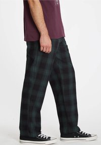 Pantalon à carreaux noirs et verts en tissu doux, avec jambes larges et coupe ample. Associé à des baskets noires et un t-shirt bordeaux.