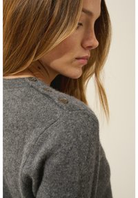 Szary sweter z dzianiny wełnianej o miękkiej fakturze, z trzema brązowymi guzikami na ramieniu i okrągłym dekoltem.