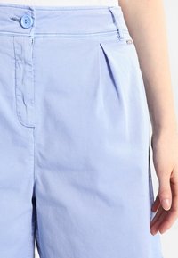 Světle modré skládané kalhoty vyrobené z jemné látky, s knoflíkovým zapínáním a bočními kapsami. Hladká textura s přizpůsobeným střihem.