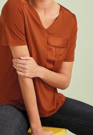 Koszulka w kolorze rdzy z dekoltem w serek, krótkimi rękawami i naszywaną kieszonką z satynowym wykończeniem po lewej stronie, zestawiona z ciemnymi dżinsami.