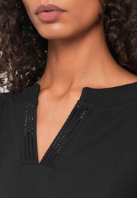 Zwarte top met een V-hals en decoratieve verticale lijnen van kleine rhinestones. Zacht stof met een gladde textuur. Close-up van de halslijn.