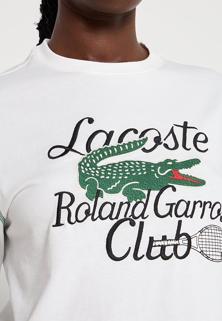Sweat-shirt blanc avec le logo crocodile vert de Lacoste et le texte "Roland Garros Club" en noir. Comprend un graphique de raquette de tennis.