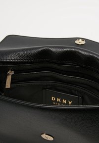 Interno di una borsa a tracolla nera in pelle, con scomparti con zip, foderato in tessuto e un'etichetta con logo dorato. Presenta una chiusura a scatto e una superficie liscia.