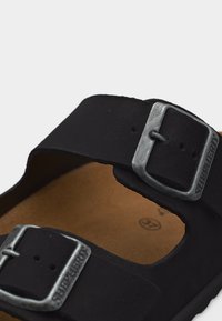 Μαύρο σανδάλι με δύο ρυθμιζόμενες μεταλλικές αγκράφες και ταμπά πέλμα, με το όνομα της μάρκας εγχαραγμένο στην αγκράφα.