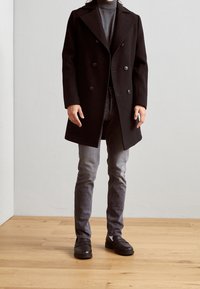 Abrigo negro de doble botonadura con solapas anchas, llevado sobre un suéter gris de cuello alto y jeans grises, completado con mocasines negros sobre un suelo de madera.