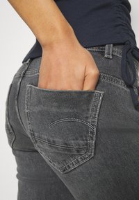 Main reposant dans la poche arrière d'un jean en denim gris foncé porté avec un haut bleu marine froncé.