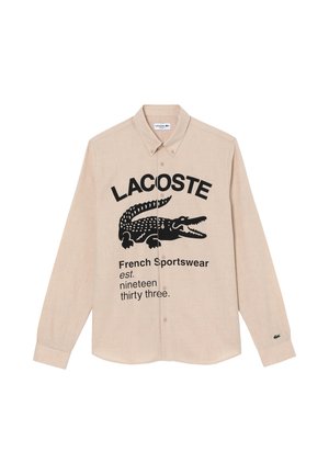 Chemise beige à manches longues boutonnée avec logo Lacoste noir, graphisme crocodile et texte "French Sportswear est. nineteen thirty three."