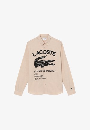 Chemise beige à manches longues boutonnée avec logo Lacoste noir, graphisme crocodile et texte "French Sportswear est. nineteen thirty three."