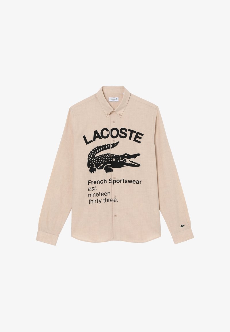 Chemise beige à manches longues boutonnée avec logo Lacoste noir, graphisme crocodile et texte "French Sportswear est. nineteen thirty three."