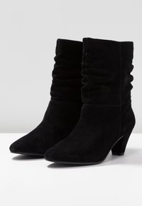 Par de botas de ante negras hasta la mitad de la pantorrilla, con cañas arrugadas, punteras puntiagudas y tacones cuadrados, sobre una superficie blanca con fondo liso.