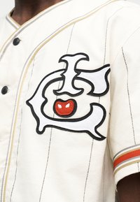 Λευκό μπέιζμπολ μπλουζάκι με κεντημένο λογότυπο σε μαύρο και λευκό με κόκκινο τόνο. Ριγέ ύφασμα στο μανίκι σε πορτοκαλί και χρυσό χρώμα.