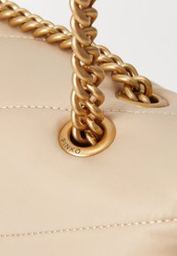 Μπεζ τσάντα tote με καπιτονέ σχέδιο, διαθέτει χρυσή αλυσίδα, μεταλλικά στοιχεία και κυκλικό λογότυπο με την επιγραφή "PINKO" αποτυπωμένο.
